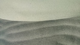Fine, textured sand forms gentle, flowing patterns in light and darker shades, creating an abstract and minimalistic visual effect.