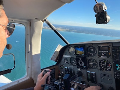 The interior of an aircraft cockpit with a pilot flying over a large body of water. The plane's instruments and controls are visible, showing various dials and screens. The view outside includes a vast expanse of blue water and a distant coastline under a clear sky.
