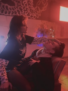 A woman with ornamental sleeve designs gently cradles the head of a person reclining on a chair in a dimly lit, atmospheric room. The setting is bathed in warm red and purple hues, with a mural or artwork adorns the wall.