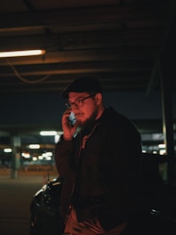 A man wearing glasses and a hat is standing in a dimly lit parking garage, holding a phone to his ear. He is dressed in a dark jacket, and the background suggests an urban setting with blurred lights and parked vehicles.