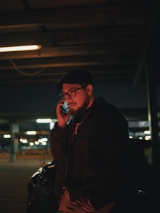A man wearing glasses and a hat is standing in a dimly lit parking garage, holding a phone to his ear. He is dressed in a dark jacket, and the background suggests an urban setting with blurred lights and parked vehicles.