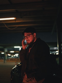 A man wearing glasses and a hat is standing in a dimly lit parking garage, holding a phone to his ear. He is dressed in a dark jacket, and the background suggests an urban setting with blurred lights and parked vehicles.
