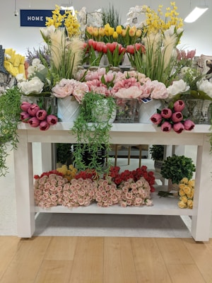 A display of various artificial flowers arranged on a white wooden table. The top shelf contains colorful tulips and lush greenery in pots, while the bottom shelf holds neatly organized bouquets of roses in pink, red, and yellow. Bright lighting enhances the vibrant colors, giving the scene a fresh and lively appearance.