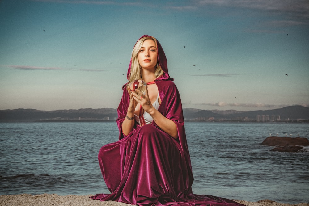 a woman in a purple dress sitting on a rock by the water