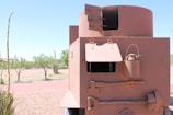 A large, rust-colored metal structure with open hatches is situated outdoors. It appears to be a part of an old, possibly military, vehicle or fortification. The structure includes a cylindrical turret and rectangular body with bolts and rivets along the edges. There are open hatches and a metal canister attached to the side, as well as tools including a large wrench and shovel. The background features a dry landscape with scattered bushy trees under a clear blue sky.