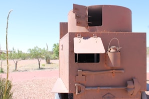 A large, rust-colored metal structure with open hatches is situated outdoors. It appears to be a part of an old, possibly military, vehicle or fortification. The structure includes a cylindrical turret and rectangular body with bolts and rivets along the edges. There are open hatches and a metal canister attached to the side, as well as tools including a large wrench and shovel. The background features a dry landscape with scattered bushy trees under a clear blue sky.