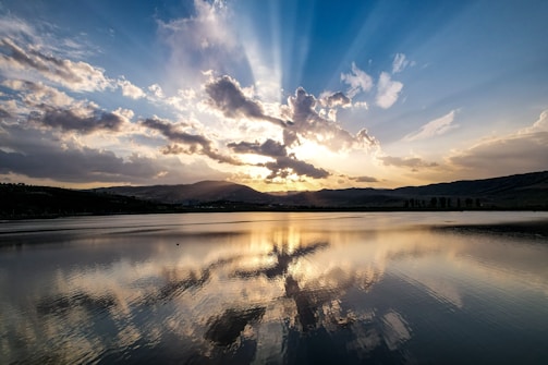 Sunset over a serene lake reflecting dramatic skies and distant mountain silhouettes.