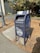 A blue metal mailbox labeled 'UC Berkeley 94720 Mail Services' stands on a paved sidewalk beside a lamppost. The mailbox has some graffiti and stickers on it, and is surrounded by dried leaves. A cyclist in a bright green shirt is visible in the background near a bike rack. Tall trees with dense foliage are also present, adding a natural element to the urban setting.