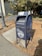 A blue metal mailbox labeled 'UC Berkeley 94720 Mail Services' stands on a paved sidewalk beside a lamppost. The mailbox has some graffiti and stickers on it, and is surrounded by dried leaves. A cyclist in a bright green shirt is visible in the background near a bike rack. Tall trees with dense foliage are also present, adding a natural element to the urban setting.