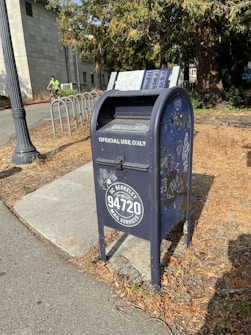 A blue metal mailbox labeled 'UC Berkeley 94720 Mail Services' stands on a paved sidewalk beside a lamppost. The mailbox has some graffiti and stickers on it, and is surrounded by dried leaves. A cyclist in a bright green shirt is visible in the background near a bike rack. Tall trees with dense foliage are also present, adding a natural element to the urban setting.
