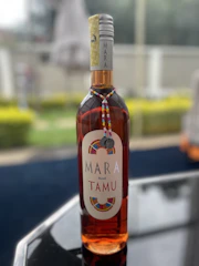 A bottle of Mara Rosé Tamu wine is placed on a reflective surface. The bottle is decorated with a colorful beaded necklace and a metallic charm. The background is blurred, featuring some greenery and possibly other garden or outdoor elements.