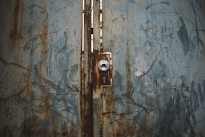 A weathered metal door with patches of rust and blue-gray paint, featuring a lock mechanism at the center.