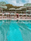 Luxurious poolside area with clear aqua blue water in the foreground and several cabanas in the background, each covered by pastel-striped umbrellas. Large, decorative leaf structures are positioned above the cabanas, and lush, green foliage serves as a backdrop.