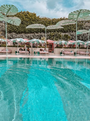 Luxurious poolside area with clear aqua blue water in the foreground and several cabanas in the background, each covered by pastel-striped umbrellas. Large, decorative leaf structures are positioned above the cabanas, and lush, green foliage serves as a backdrop.