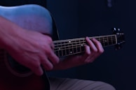 Hands are positioned on the fretboard of an acoustic guitar, with fingers pressing down on the strings against a dark, subdued background. The scene is dimly lit with a cool-toned ambiance.