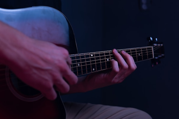 Hands are positioned on the fretboard of an acoustic guitar, with fingers pressing down on the strings against a dark, subdued background. The scene is dimly lit with a cool-toned ambiance.