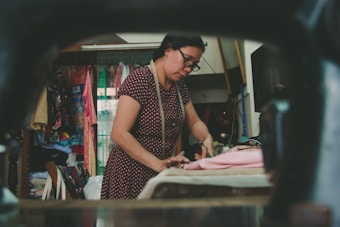 A woman is working on fabric in a sewing room. She appears focused, wearing glasses and a patterned dress, with a measuring tape around her neck. The room is filled with colorful fabrics and sewing materials, indicating a workshop or tailoring environment.