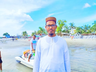A man in a white traditional outfit and a patterned cap stands on a beach with palm trees and buildings in the background. The shoreline is bustling with people, some walking by and others engaged in activities. A person is seen sitting on a jet ski in the water.