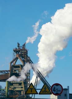 A large industrial structure with pipes and smokestacks, emitting white smoke into a bright blue sky. In the foreground, several road signs are visible, including a pedestrian crossing sign and a height restriction sign.