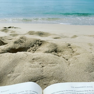 A serene beach scene with gentle waves lapping at the shore. The foreground features a book open on the sand, suggesting relaxation and leisure.