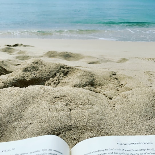 A serene beach scene with gentle waves lapping at the shore. The foreground features a book open on the sand, suggesting relaxation and leisure.