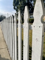 A white wooden picket fence lines a sidewalk on a sunny day, with a house partially visible in the background. Tall, leafy trees create a shady atmosphere, offering a stark contrast to the bright blue sky above. The fence features a classic pointed design, and the green grass on the other side adds a natural touch to the suburban scenery.