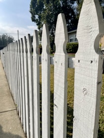 A white wooden picket fence lines a sidewalk on a sunny day, with a house partially visible in the background. Tall, leafy trees create a shady atmosphere, offering a stark contrast to the bright blue sky above. The fence features a classic pointed design, and the green grass on the other side adds a natural touch to the suburban scenery.