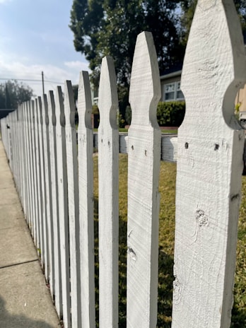 A white wooden picket fence lines a sidewalk on a sunny day, with a house partially visible in the background. Tall, leafy trees create a shady atmosphere, offering a stark contrast to the bright blue sky above. The fence features a classic pointed design, and the green grass on the other side adds a natural touch to the suburban scenery.