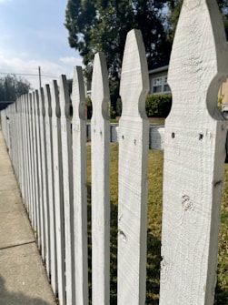 A white wooden picket fence lines a sidewalk on a sunny day, with a house partially visible in the background. Tall, leafy trees create a shady atmosphere, offering a stark contrast to the bright blue sky above. The fence features a classic pointed design, and the green grass on the other side adds a natural touch to the suburban scenery.