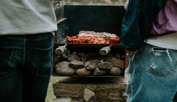two people standing next to a grill with food on it