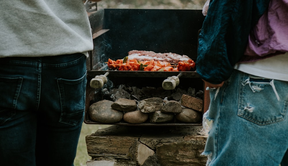 two people standing next to a grill with food on it