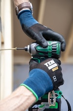 A person wearing blue and black protective gloves is using a green and black cordless drill to insert a screw into wood. The person is wearing a brown watch on their wrist. The drill has branding labels visible.
