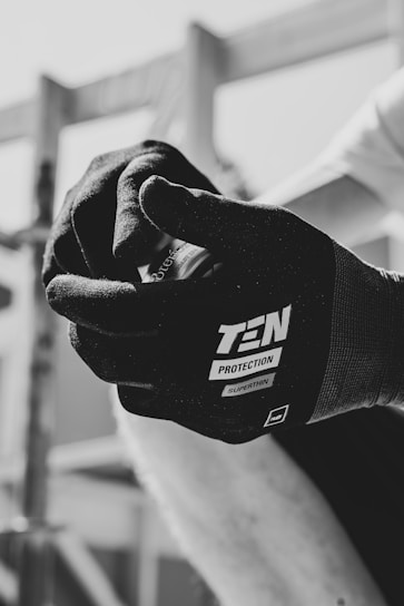 A close-up in black and white captures a hand wearing a glove labeled 'TEN Protection Superthin.' The hand is holding an object, possibly a can, with a blurred background indicating an outdoor or construction setting.