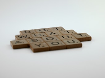Wooden tiles with letters arranged to spell 'BUT WHAT ABOUT YOU', resembling Scrabble pieces. The background is a smooth gradient of white to light gray, creating a soft and minimalist look.