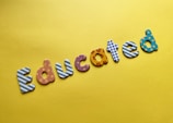 Colorful letters spelling the word 'Educated' are arranged on a bright yellow background. Each letter features a different pattern, such as stripes, polka dots, flowers, and stars.