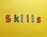 Colorful, patterned letters spelling out 'SKILLS' are arranged on a bright yellow background. Each letter features distinct patterns: red with white hearts, black with white stars, purple with yellow stars, and green with pink polka dots.