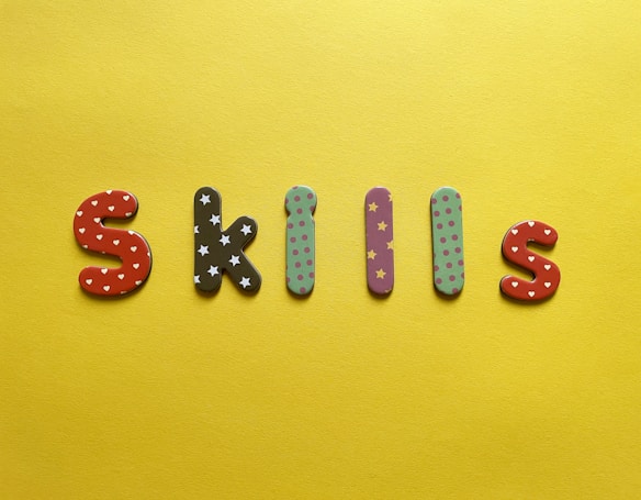 Colorful, patterned letters spelling out 'SKILLS' are arranged on a bright yellow background. Each letter features distinct patterns: red with white hearts, black with white stars, purple with yellow stars, and green with pink polka dots.