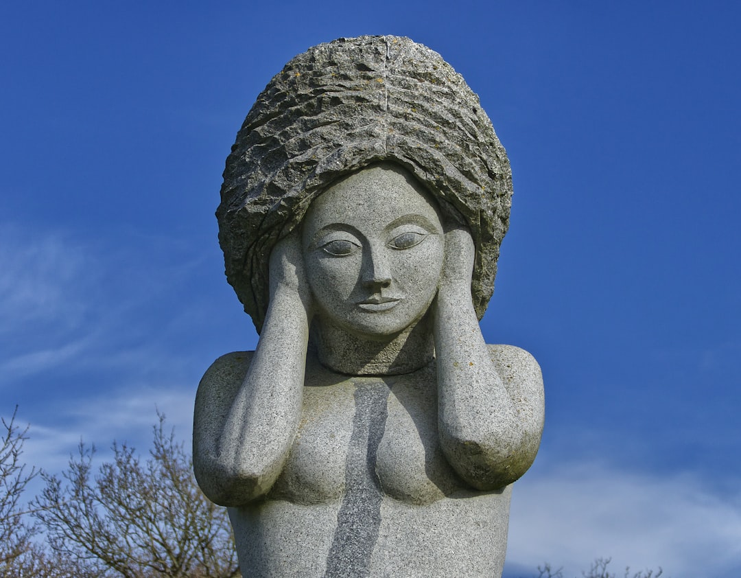 a statue of a woman holding her head with her hands, La sculpture en granit « Eodez » réalisée par l’artiste Didier Hardellet assisté de Cécile Maurage.<p></figure>Eodez est la soeur de Tanguy, canonisé saint Tanguy de Locmazhé, et fille du seigneur de Trémazan au VIe siècle. Tanguy, en 545, lui coupa la tête après avoir eu vent de calomnies propagées par leur belle mère. Après que Tanguy ait avoué son méfait à son père, Eodez réapparut et remit sa tête sur son tronc. Elle pardonna à son frère et mourut en sainte femme.