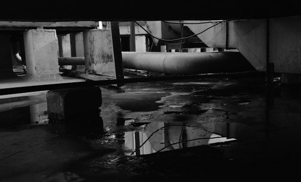 A dark, industrial environment with visible pipes, concrete supports, and reflective puddles of water on the ground. The lighting is minimal, adding a moody and gritty atmosphere to the scene.