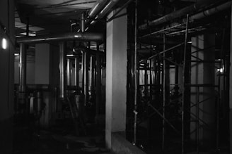 A dark, industrial setting featuring a network of metal pipes and scaffolding within a confined space. The lighting is dim, casting shadows across the pipes and creating a mysterious atmosphere.