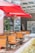 Bright red umbrellas provide shade for an outdoor patio area with wooden chairs and tables. Glass windows reflect the sun, while green foliage adds a natural touch. A 'no parking' sign is visible, along with directional signs in the background.