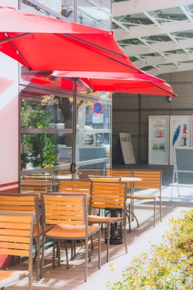 Bright red umbrellas provide shade for an outdoor patio area with wooden chairs and tables. Glass windows reflect the sun, while green foliage adds a natural touch. A 'no parking' sign is visible, along with directional signs in the background.