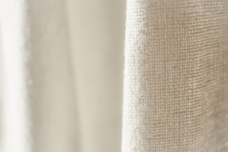 A close-up view of a textured fabric with a light cream color. The weave pattern of the textile is visible, highlighting the natural fibers and intricate details.
