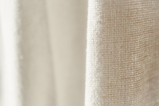 A close-up view of a textured fabric with a light cream color. The weave pattern of the textile is visible, highlighting the natural fibers and intricate details.