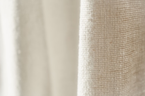 A close-up view of a textured fabric with a light cream color. The weave pattern of the textile is visible, highlighting the natural fibers and intricate details.