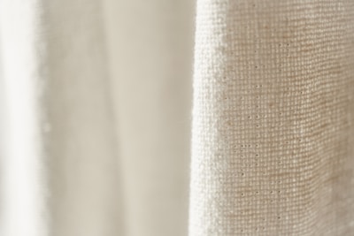 A close-up view of a textured fabric with a light cream color. The weave pattern of the textile is visible, highlighting the natural fibers and intricate details.