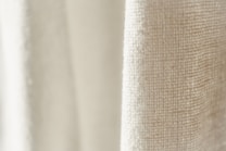 A close-up view of a textured fabric with a light cream color. The weave pattern of the textile is visible, highlighting the natural fibers and intricate details.