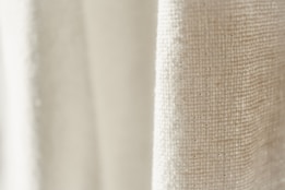 A close-up view of a textured fabric with a light cream color. The weave pattern of the textile is visible, highlighting the natural fibers and intricate details.