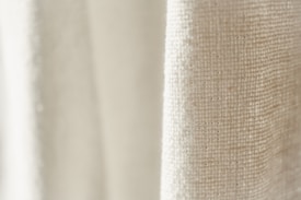 A close-up view of a textured fabric with a light cream color. The weave pattern of the textile is visible, highlighting the natural fibers and intricate details.