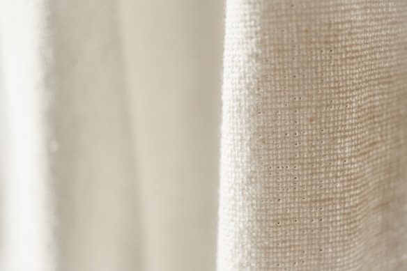 A close-up view of a textured fabric with a light cream color. The weave pattern of the textile is visible, highlighting the natural fibers and intricate details.